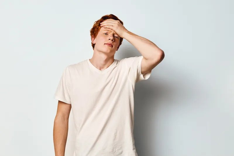 Young man in casual white t shirt with red hair standing against plain wall holding forehead with closed eyes showing tired stressed emotion and discomfort indoors.