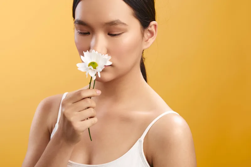 asian woman holds a white flower near her nose, eyes closed, in a soft studio setting with warm lighting, evoking calm, beauty, and gentle serenity.