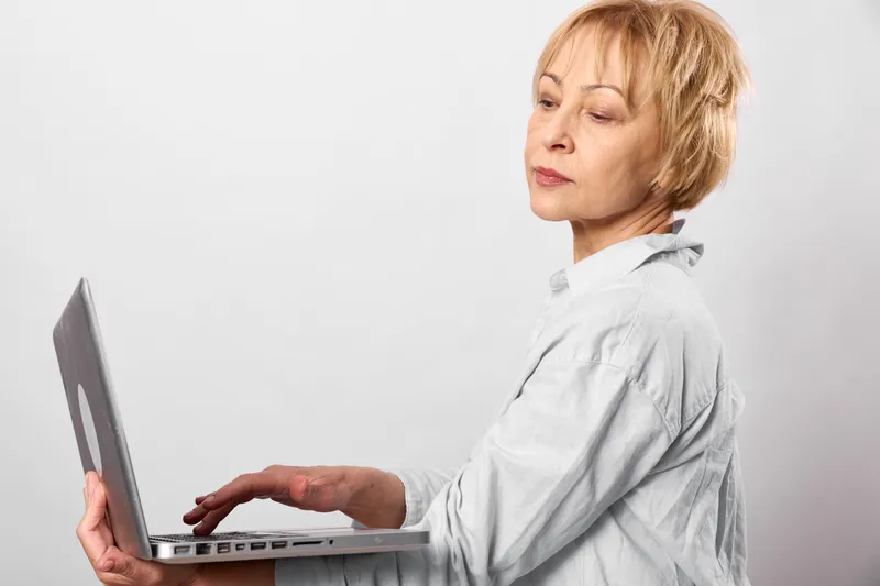Woman with short blonde hair using a laptop in office setting, side view, focused expression, hands on keyboard, white shirt, light, minimal background