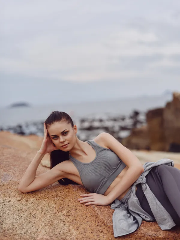 Young woman in athletic wear posing on a rocky beach backdrop, displaying relaxed emotion and natural beauty, highlighting fitness and wellness concepts