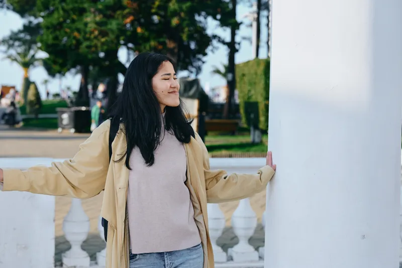 Woman outdoor pose standing beside a column in a park, wearing a beige coat and sweater, sunny daylight, relaxed mood, urban architecture and nature in view against bright sky view