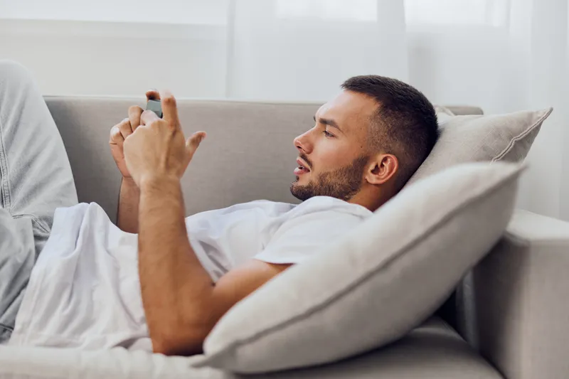 Relaxed man using smartphone while lying on the sofa, showcasing modern home living. A cozy atmosphere enhances a sense of leisure and connection in everyday life.