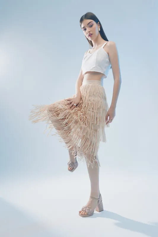 Slim woman in a white crop top and fringed beige skirt poses with one leg raised on a light background. Concept of woman beauty fashion and modern style.
