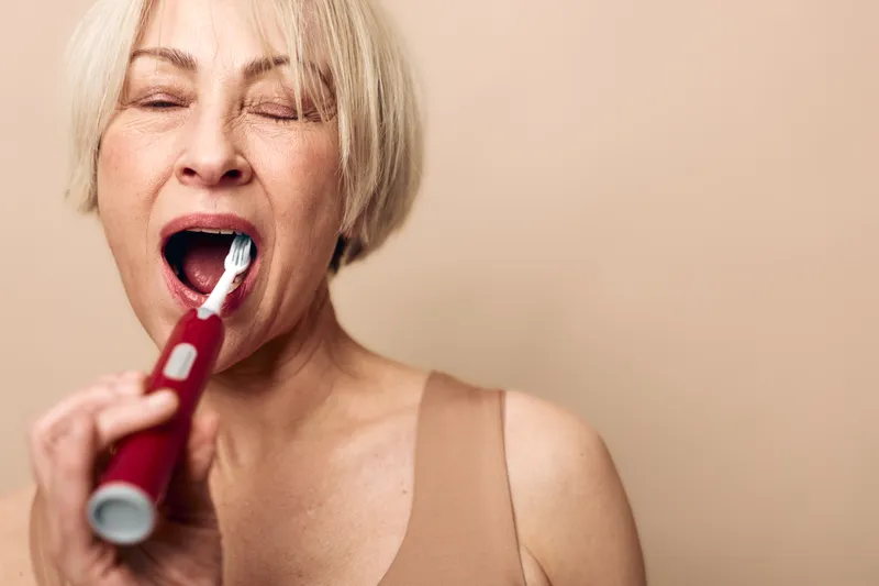 Mature woman brushing teeth with electric toothbrush, eyes closed, enjoying oral care routine in beige room. Concept of dental hygiene, self-care, and healthy lifestyle for seniors.
