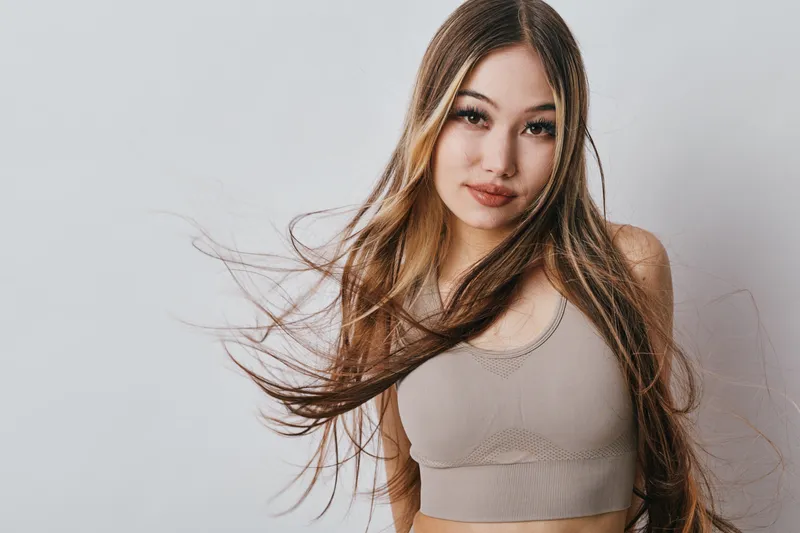 Young woman with long hair wearing cropped top standing against plain background. Female model looking at camera with natural makeup and flowing hair movement.