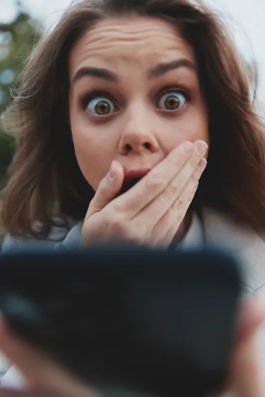 Surprised woman with wide eyes covering mouth looking at smartphone, emotional reaction, casual clothing, outdoor natural background