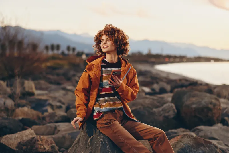 Woman lifestyle portrait on rocky beach coast at sunset, curly haired woman in orange jacket holding smartphone, relaxed sitting pose, golden light and mountains in distance, travel mood