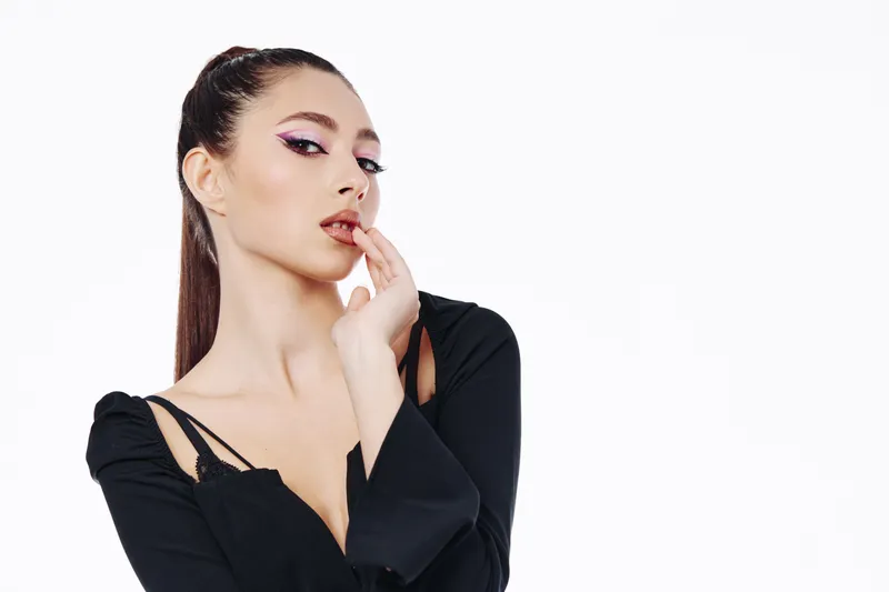 Fashionable woman with sleek hair in a modern black outfit poses with a pensive expression, showcasing bold eye makeup and clean styling on white background. Concept of beauty and fashion.