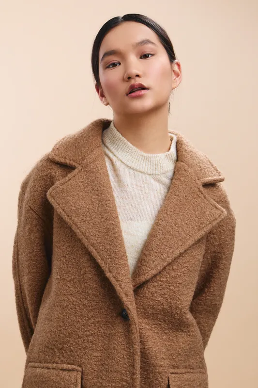 Asian woman stands in a neutral studio, wearing a camel wool coat and knit sweater, exuding calm confidence and refined style in a minimalist fashion portrait.