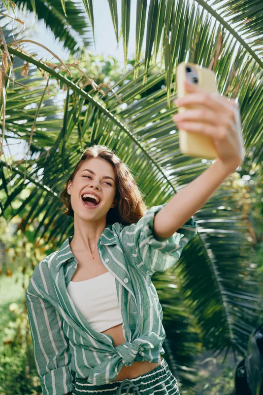 Cheerful woman selfies in a sunny tropical setting, wearing a striped green shirt over a white top, arms extended, bright smile, vibrant energy.