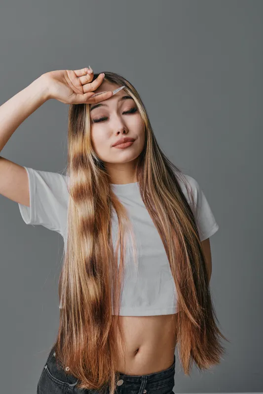 Woman with long straight hair in casual white cropped t shirt posing with eyes closed and hand on forehead against gray background indoor portrait, fashion style.