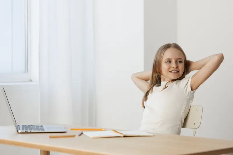 Cheerful young girl relaxing at desk with laptop, enjoying study time, minimalistic background, bright white tones, casual outfit reflecting confidence