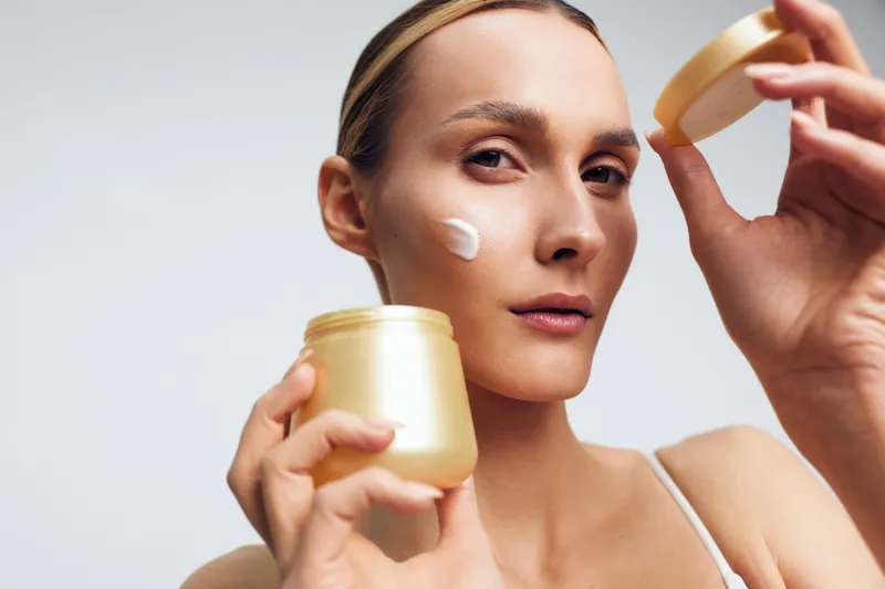 Young woman applies moisturizing cream on face holds jar and lid closeup skincare routine natural beauty care isolated on white background with serious expression.