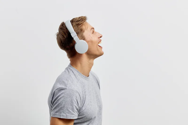 Smiling young caucasian man wearing white headphones and casual gray t-shirt, looking upwards with joy and enthusiasm, isolated on plain light background, studio portrait. People lifestyle concept