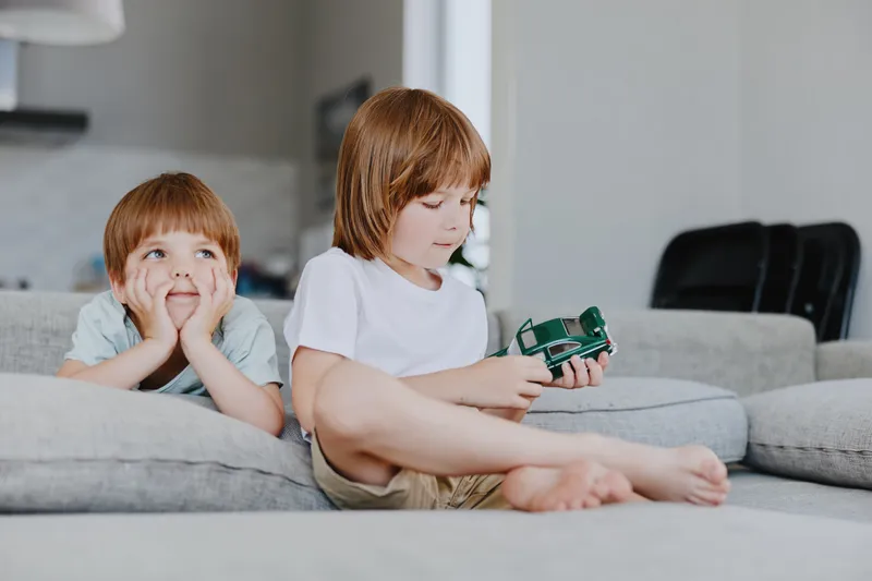 Two children enjoying playtime on a cozy sofa, one boy is focused on a toy car while the other gazes thoughtfully. A fun moment of childhood joy and creativity.