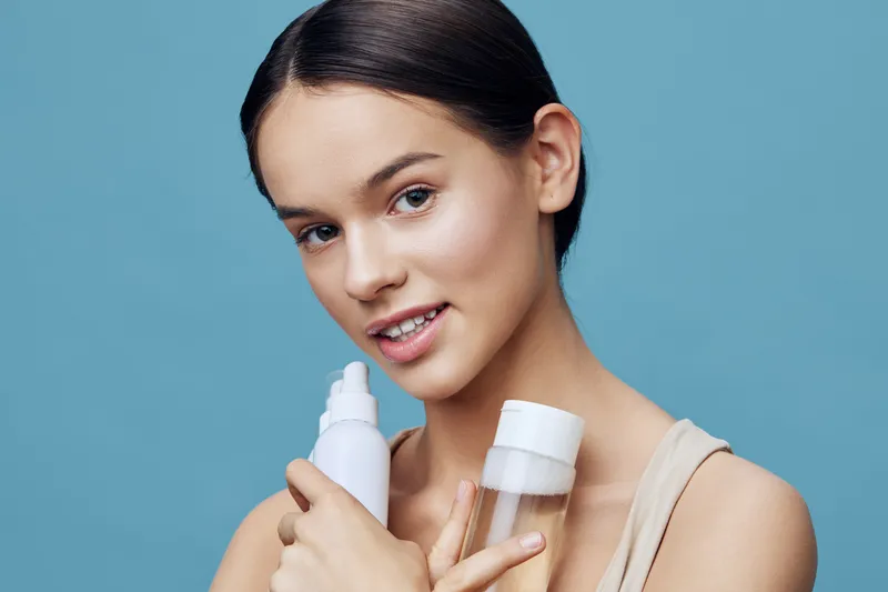 Young model holding skincare products, promoting healthy beauty on a soft blue background, showcasing natural glow and confidence