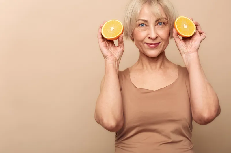 Smiling mature woman with short blonde hair holds two orange halves near face on beige background. Concept of healthy lifestyle, natural vitamins, skincare, and cheerful positive mood.