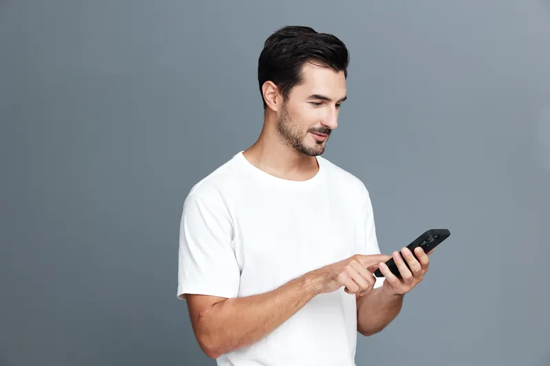 Handsome smiling young man using a mobile phone while standing against a plain gray background. This portrait captures modern lifestyle and technology interaction.