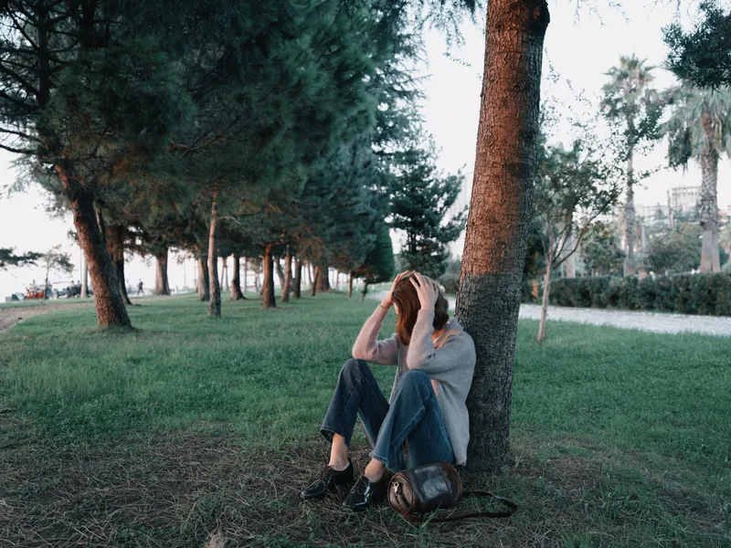 A solitary figure rests beneath a tree in a quiet park, leaning against the trunk. The mood is pensive and calm, suggesting reflection, fatigue, and a pause from daily routines in a green setting.