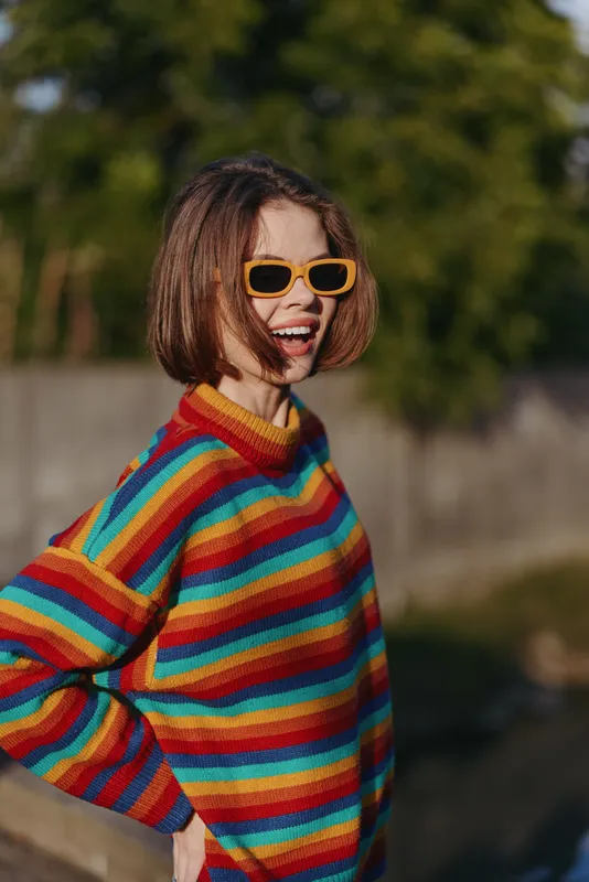Woman smile portrait outdoors in colorful striped sweater and yellow sunglasses, casual fashion look with short bob hair, joyful expression and warm natural light.