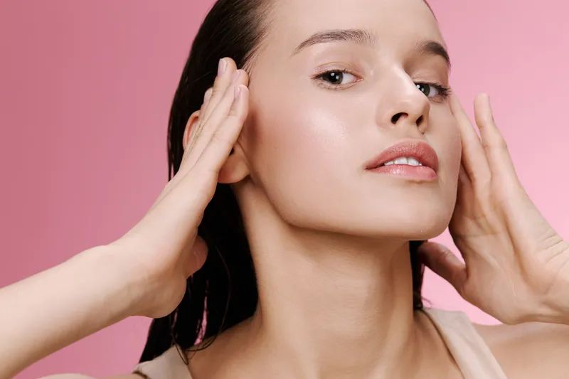 Young woman applying skincare routines with a calm expression on a soft pink background emphasizing beauty and self care