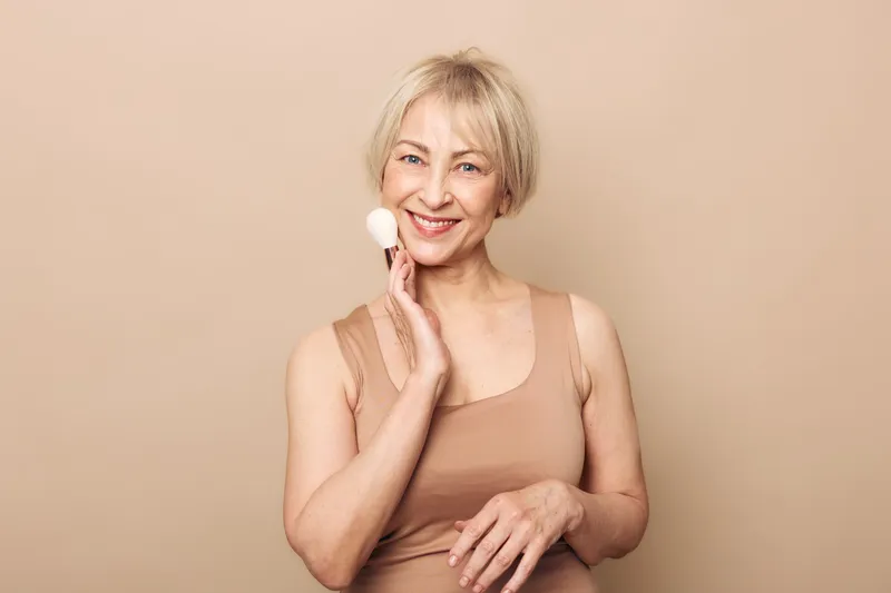 Smiling mature woman with short blonde hair applying makeup powder on her cheek with a brush against beige background. Concept of beauty, skincare, and natural aging.