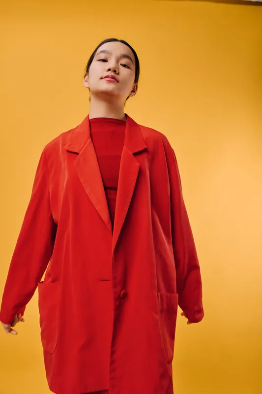 Young woman wearing oversized red coat and matching sweater posing confidently against orange background in studio. Fashion, model, casual style, portrait with modern urban look and attitude.