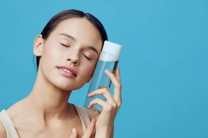 Young woman with closed eyes holding a skincare product, conveying serenity and beauty against a bright blue background for wellness and self care
