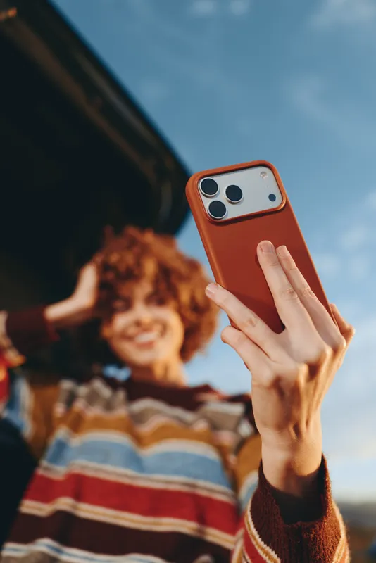 Woman hand holds smartphone for selfie outdoors with orange case, background shows smiling child with curly hair softly blurred, bright daylight sky, casual lifestyle portrait concept