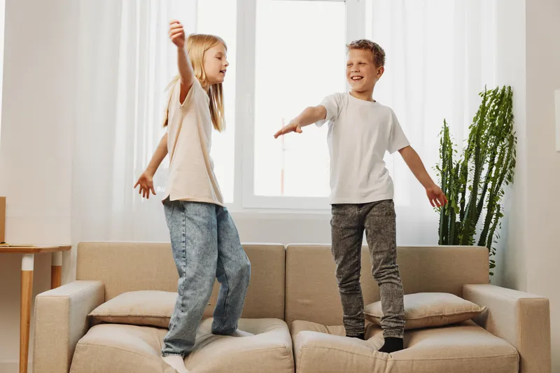 Two happy children jumping on a beige sofa in a bright living room with natural light and green plant. Concept of playful kids, home fun, and joyful family moments.
