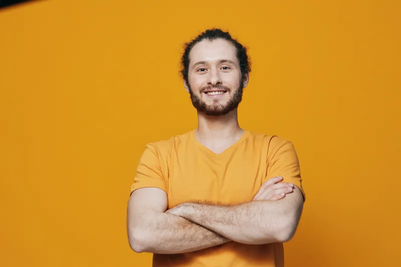 Cheerful young man with a beard in an orange t shirt smiling against a vibrant yellow background, showcasing confidence and positivity