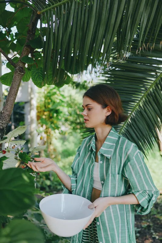 A woman in a striped green shirt tends plants outdoors, holding a white bowl as tropical foliage frames the scene.