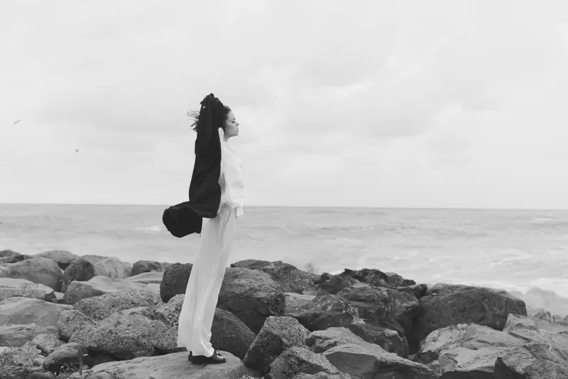 Silhouette on rocky coastline, wind swirling dress, calm ocean and cloudy sky, a contemplative stance by the shore
