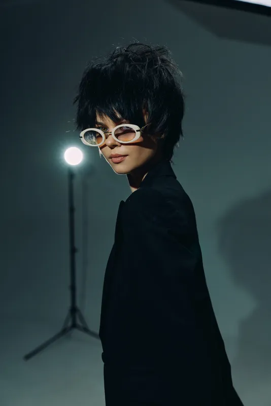 Stylish woman wearing white sunglasses and black outfit posing in studio with soft lighting and dark background.