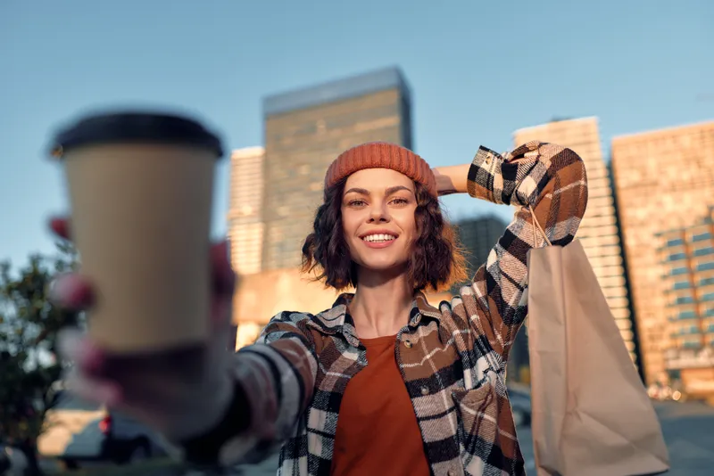 Woman holding coffee cup and shopping bag in city, smiling candidly in beanie and plaid, urban golden hour glow and authenticity, candid lifestyle, mindful living, emotional storytelling.