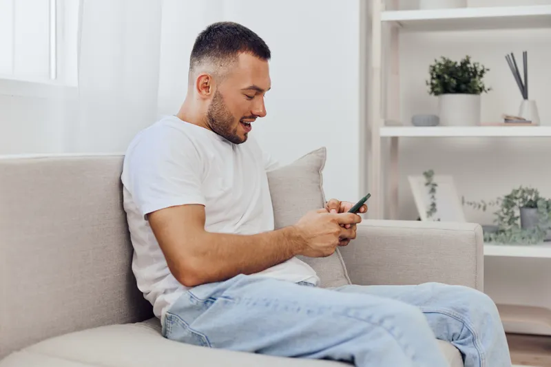Engaged man using smartphone on a cozy sofa, wearing casual clothing, surrounded by modern decor, illustrating convenience and relaxation in digital life.