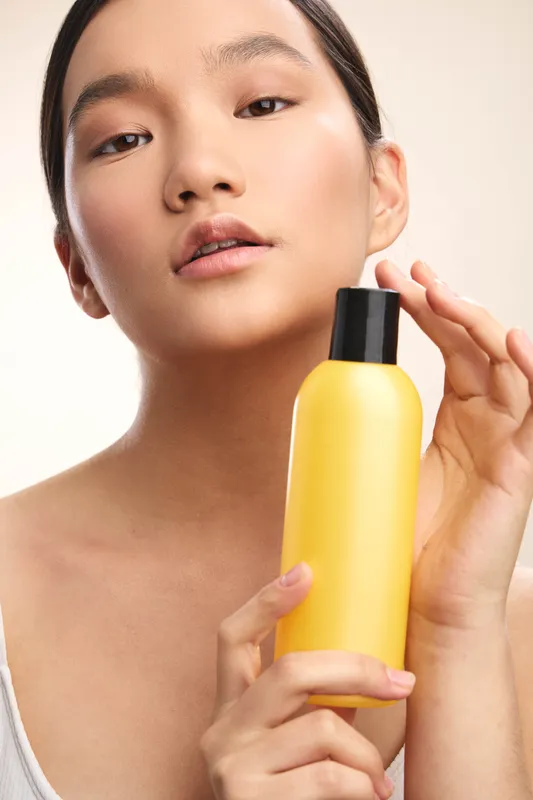 asian woman presents a bright yellow bottle close to her face, showcasing skincare product with smooth skin, soft lighting, and a calm, confident mood in a clean backdrop.