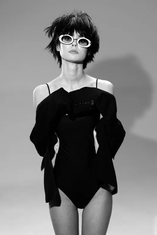 Black and white portrait of a stylish woman wearing sunglasses and a fashionable black outfit with short hair and confident pose.