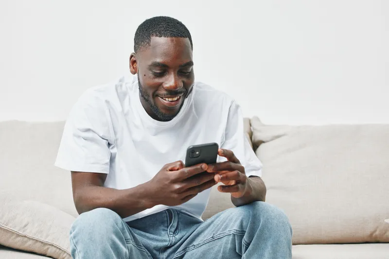 Happy young African man smiling while using a smartphone at home, wearing casual clothing, relaxed atmosphere and neutral background