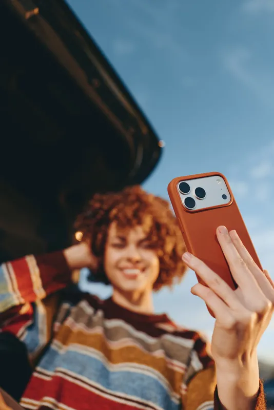 Woman lifestyle, smartphone selfie in hand, smiling woman portrait with curly hair outdoors under blue sky, casual plaid sweater, communication and technology, soft blurred background