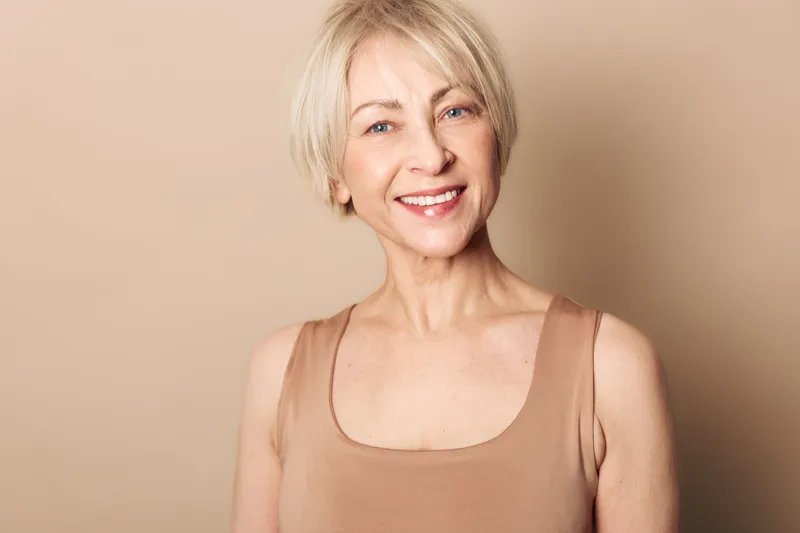 Smiling mature woman with short blonde hair wearing beige tank top poses against a soft neutral background. Concept of natural beauty, confidence, aging, and healthy lifestyle.