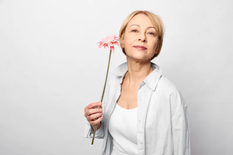Woman holding flower in studio portrait, mature calm expression, soft light on face, pastel pink bloom, white background, relaxed posture, spring mood, quiet wellbeing concept