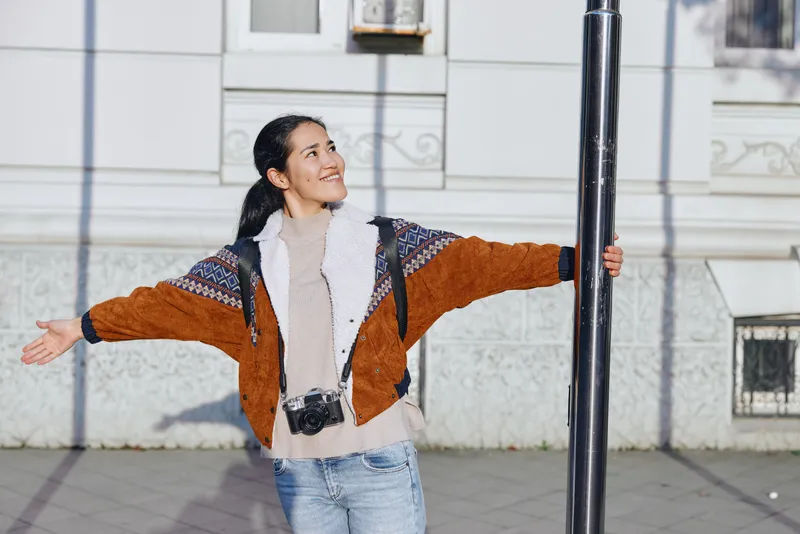 Outdoor street scene with a smiling woman spreading arms beside a pole, wearing a warm brown jacket and jeans, a joyful candid moment ideal for lifestyle travel and fashion stock