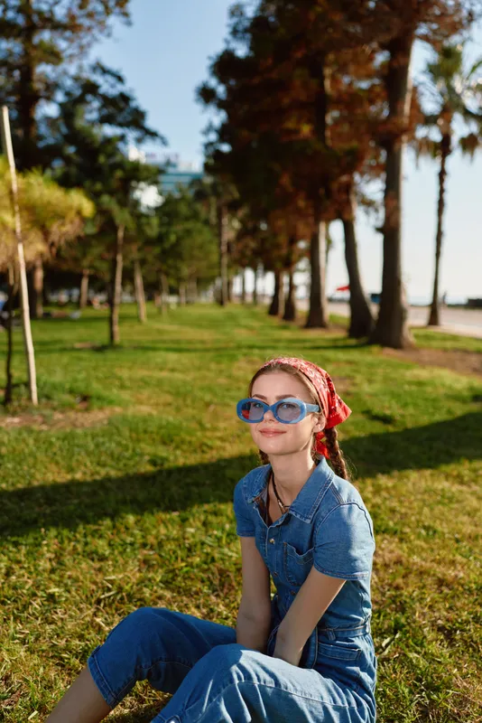 Lifestyle woman in modern boho-western denim street style sitting on grass wearing blue sunglasses with film color effect and a red bandana headscarf outdoors.