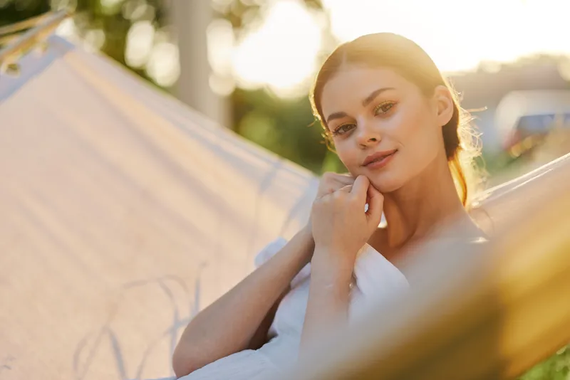 Young woman relaxing in a hammock at sunset, showcasing a serene expression in a natural setting, wearing a light outfit, evoking tranquility and peace