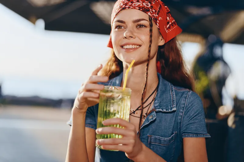 Lifestyle woman in modern boho-western denim street style enjoying refreshing drink, film color tones, smiling with relaxed summer mood and stylish accessories.