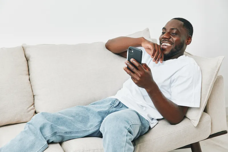 Young African man laughing while using smartphone on a comfortable couch in a modern living room, showcasing joy and relaxation