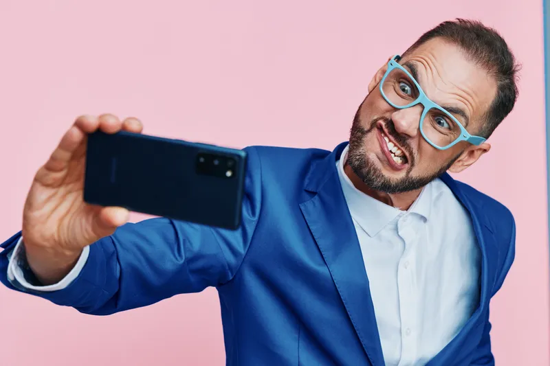 A focused male professional stands before a solid colored backdrop, taking a confident selfie in a smart suit and stylish glasses, signaling optimism and leadership in a modern corporate portrait.