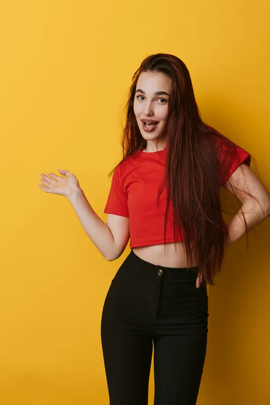 Young woman with long hair in red crop top and black pants posing with hand raised and smiling on yellow background for lifestyle and fashion portrait.