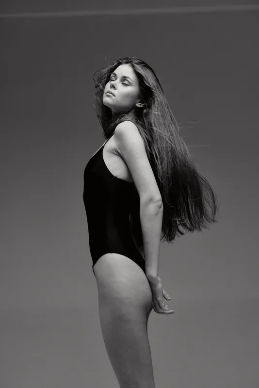 Black and white portrait of a young woman in a black swimsuit with long hair, posing confidently with closed eyes against plain background.